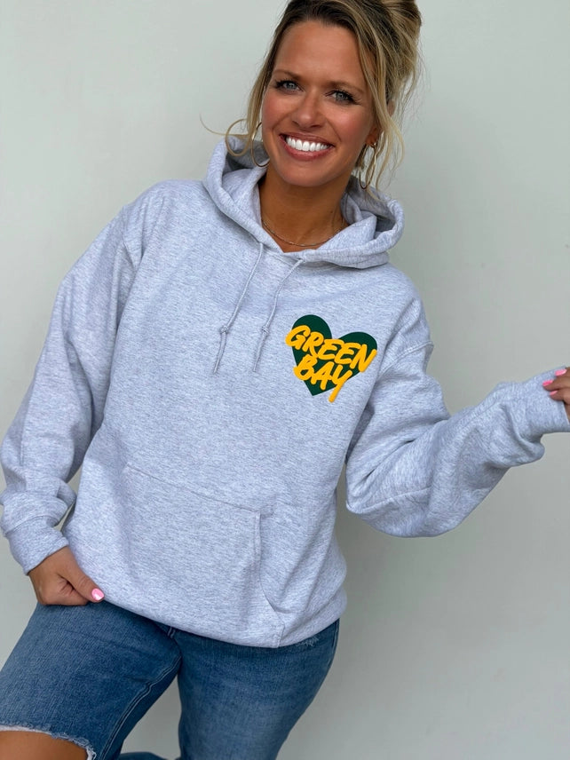 Model wearing an ash gray hoodie with a green and gold Green Bay heart puff print on the chest, front view