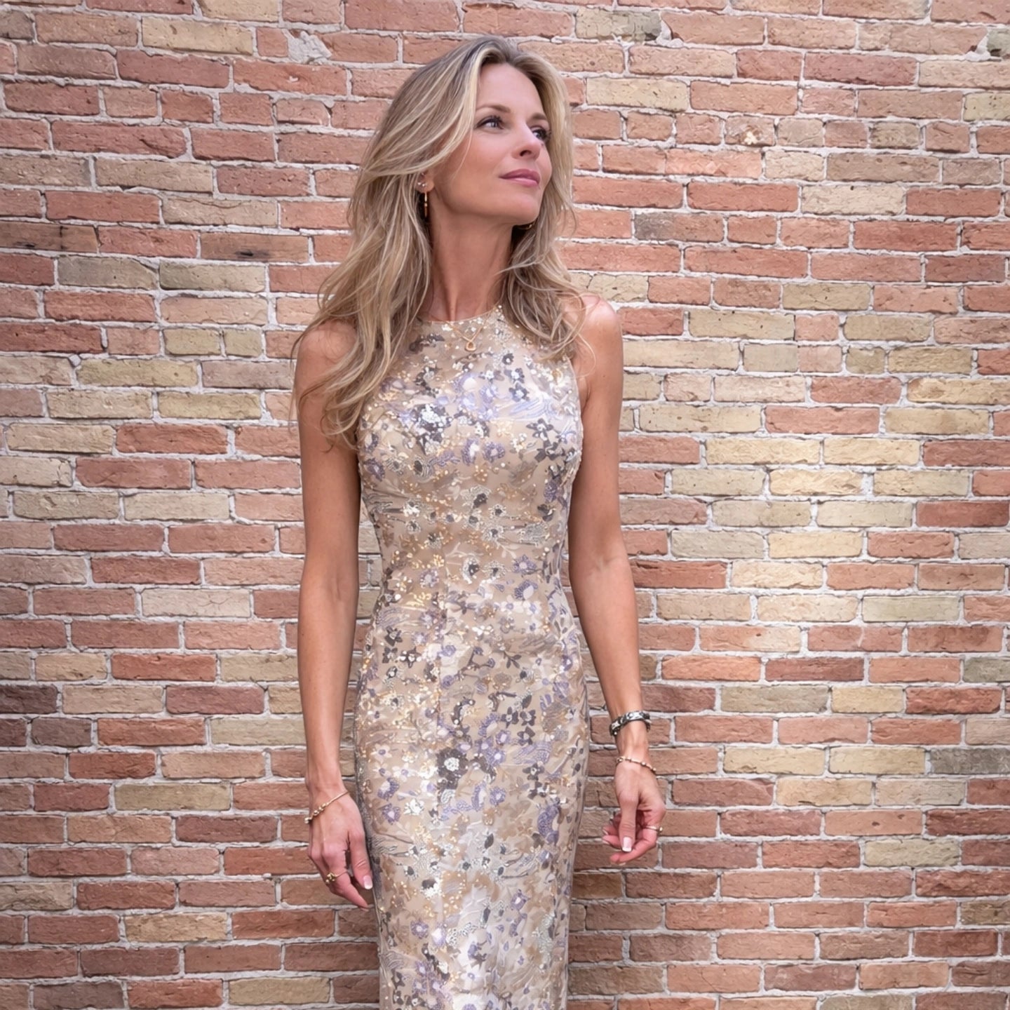 Model wearing a sleeveless champagne and silver floral sequin midi dress against a brick wall